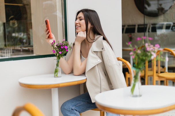 счастливая женщина с длинными волосами сидит на стуле возле бистро стола с цветами в вазе и смс на смартфоне, сидя в модной одежде с бежевым блейзером в кафе на террасе в Стамбуле  - Фото, изображение