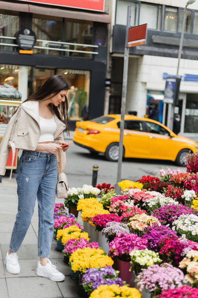 brunette vrouw met lang haar staande in beige leren jas, spijkerbroek en handtas met ketting riem houden smartphone in de buurt van bloemen naast wazig straat in Istanbul, verkoper - Foto, afbeelding