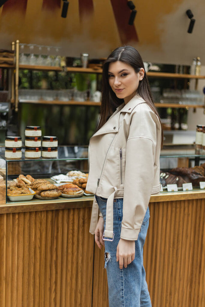brunette young woman with long hair in beige leather jacket and denim jeans looking at camera while standing near cake display with tasty pastry and jars of jam in modern bakery shop in Istanbul  - Fotografie, Obrázek