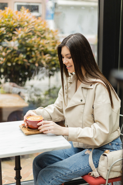 šťastná brunetka žena v kožené bundě sedí na židli vedle okna a bistro stůl, zatímco drží šálek cappuccino s kávou art uvnitř moderní kavárny v Istanbulu  - Fotografie, Obrázek