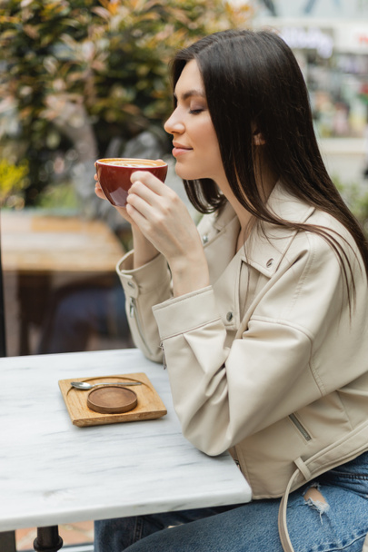 szczęśliwa brunetka kobieta z zamkniętymi oczami w skórzanej kurtce siedzi na krześle obok okna i stołu bistro trzymając filiżankę cappuccino wewnątrz nowoczesnej kawiarni w Stambule  - Zdjęcie, obraz