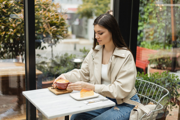 mulher morena com cabelos longos sentados em jaqueta de couro ao lado da janela e da mesa bistrô enquanto segura xícara de cappuccino com arte do café perto de cheesecake dentro do café moderno em Istambul  - Foto, Imagem