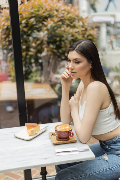 jonge vrouw met lang haar zit naast het raam en bistro tafel met kopje cappuccino en smakelijke cheesecake op bord, terwijl weg te kijken in het moderne cafe in Istanbul  - Foto, afbeelding