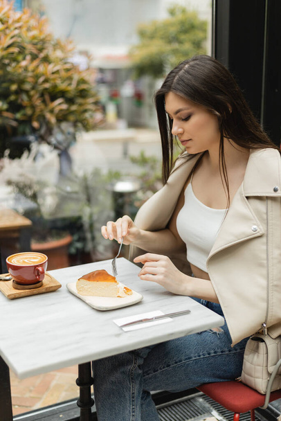 イスタンブールのモダンなカフェの窓の近くの革のジャケットに座っている間、若いブルネットの女性はビストロテーブルの上にコーヒーアートとカプチーノのカップの横にチーズケーキの近くにフォークを持っている  - 写真・画像