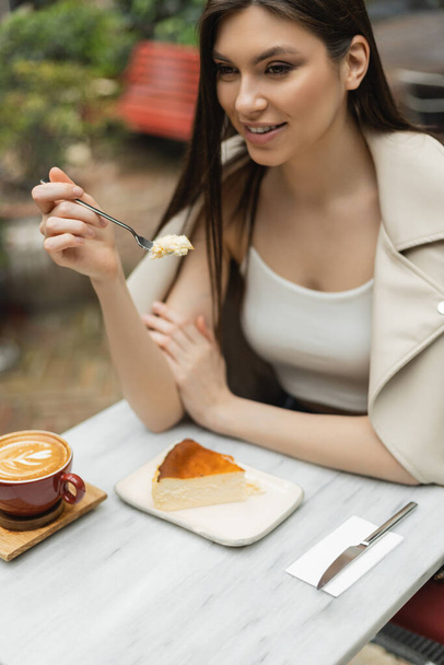 mulher sorridente com garfo de cabelo comprido segurando com cheesecake ao lado da xícara de cappuccino com arte do café na mesa bistrô enquanto sentado dentro do café moderno em Istambul  - Foto, Imagem