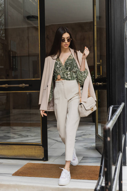 brunette and young woman in trendy outfit and sunglasses walking out of building while holding handbag with chain strap on modern street in Istanbul, entrance door  - Photo, Image