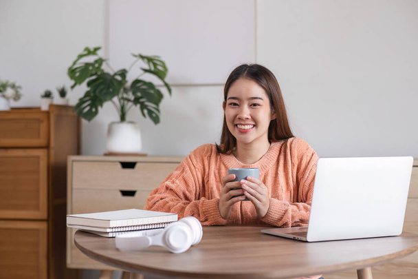 Kaunis nuori hymyilevä nainen työskentelee nauttien kannettavasta tietokoneesta ja juoden kahvia istuen mukavasti kotona. - Valokuva, kuva