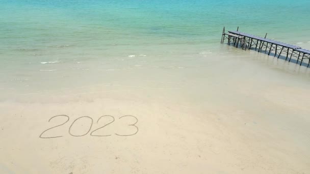 Luchtfoto van het woord 2023 geschreven op het witte zand op een tropisch strand met turquoise oceaanwater, en een houten brug op de achtergrond. - Video