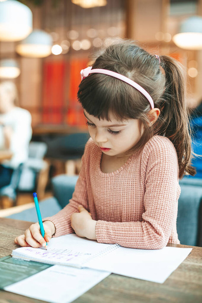 Roztomilá holčička píše něco do sešitu sedící v kavárně - Fotografie, Obrázek