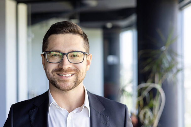 Close-up photo portrait of young successful entrepreneur, businessman investor wearing glasses at workplace smiling and looking at camera. - Fotografie, Obrázek