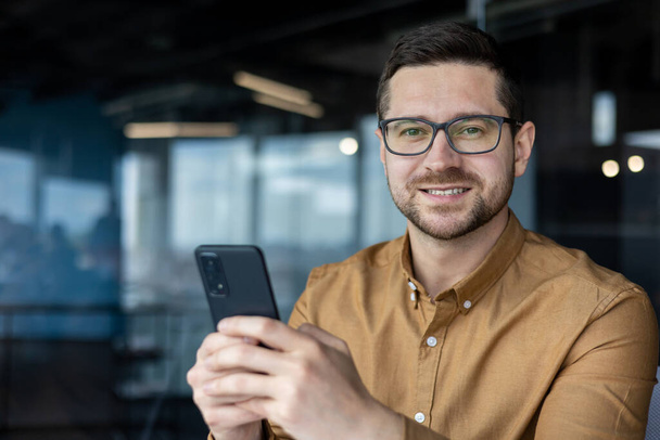 Closeup portrait of a programmer with a phone, a man in a shirt at the workplace smiles and looks at the camera, a businessman holds a phone, uses an online application, makes a call, and browses. - Foto, afbeelding
