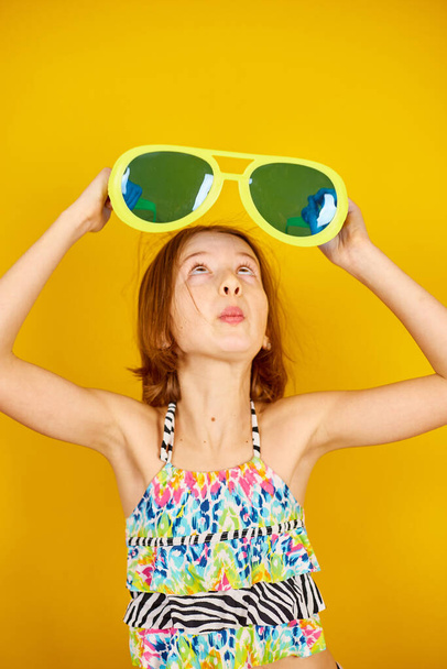 Улыбчивая девочка-подросток в купальнике и больших солнцезащитных очках позирует, весело, делает лица в студии на желтом фоне, летнее настроение, - Фото, изображение