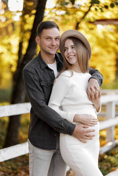 Happy stylish pregnant woman and man hugs, kiss, touches belly and listen baby's kicks, walk outdoor in autumn park. Pregnancy, motherhood and parenthood concept. Family is expecting for unborn baby - Zdjęcie, obraz
