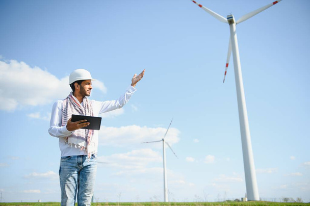 Engineer India man working at windmill farm Generating electricity clean energy. Wind turbine farm generator by alternative green energy. Asian engineer checking control electric power. - Photo, image