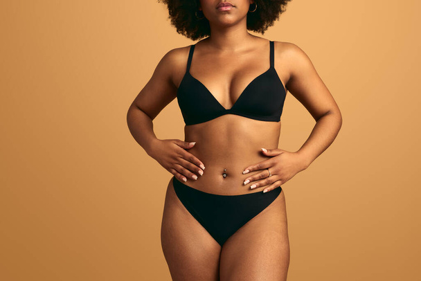 Crop anonymous serious ethnic female plus size model in black underwear with curly dark hair touching belly against beige background - Φωτογραφία, εικόνα
