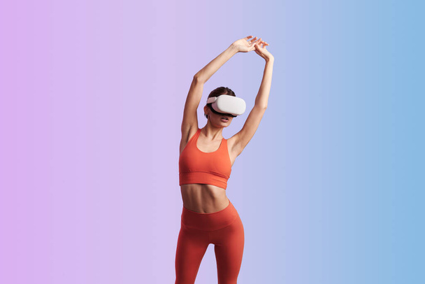 Slim lady in VR goggles wearing red crop top and leggings while doing exercises with raised arms against gradient background - 写真・画像