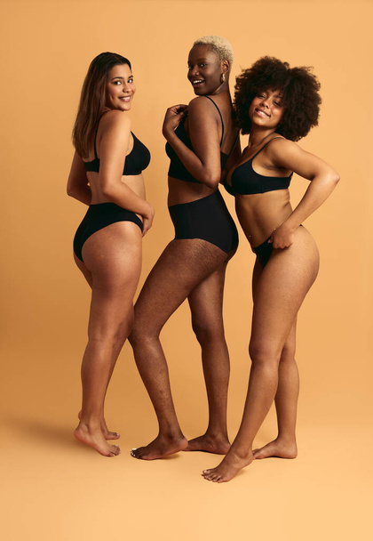 Full body of young multiracial girlfriends in black lingerie standing against orange background smiling and looking at camera - Photo, Image