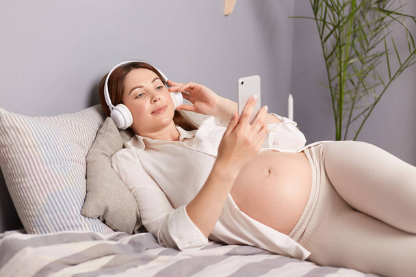 Expecting female with stunning appearance listening to music with headphones and smartphone, feeling the rhythm while revealing her tummy in a comfy outfit. - Foto, imagen