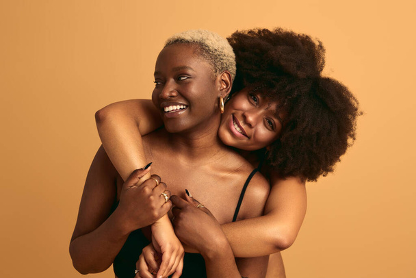 Happy African female with Afro hair embracing girlfriend from behind while standing against orange background having fun - Foto, Imagem