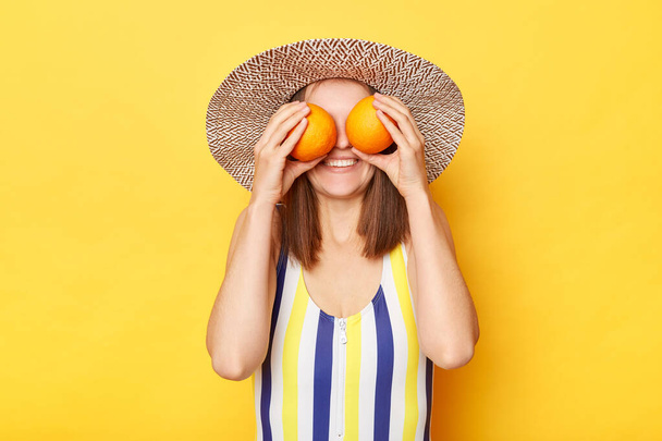 Unrecognizable smiling woman wearing striped swimming suit and sun hat isolated on yellow background hiding her eyes behind oranges enjoying tropical rest. - 写真・画像