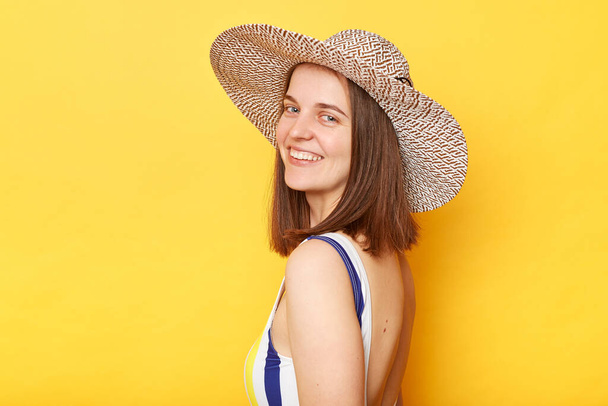 Side view portrait of smiling woman wearing striped swimming suit and sun hat isolated on yellow background looking at camera with happy face, relaxing on beach, resting. - Fotografie, Obrázek