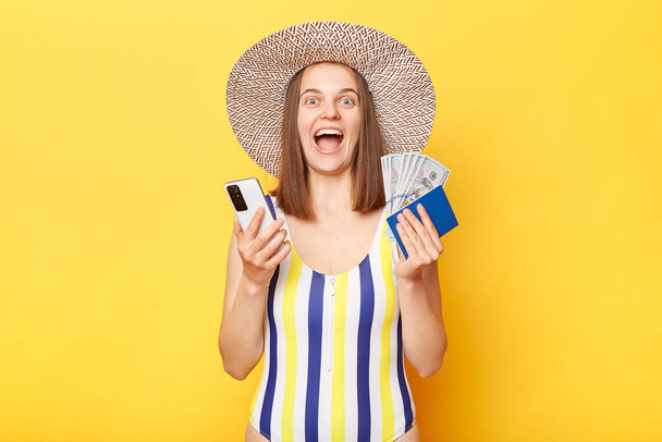 Optimistic smiling woman on resort dressed in striped swimwear and straw hat posing isolated over yellow background holding her passport and using smart phone yelling with happy face. - Φωτογραφία, εικόνα