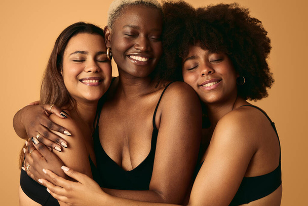 Group of cheerful multiracial female friends in black lingerie smiling and hugging each other with closed eyes against beige background in studio - Foto, Imagen