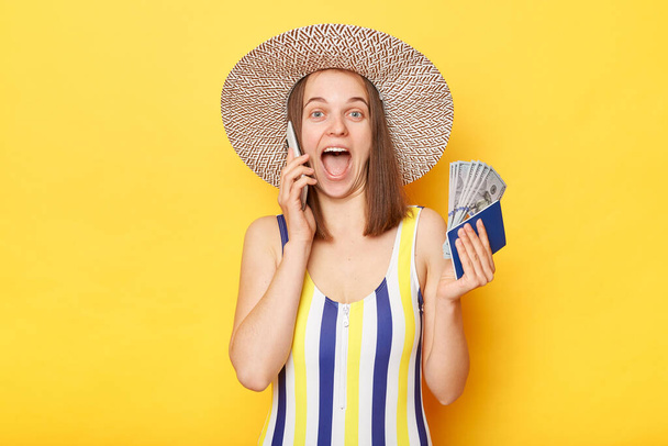 Unexpected vacation. Happy amazed woman on resort dressed in striped swimwear and straw hat posing isolated over yellow background holding passport and money talking phone boasting her vacation. - Fotoğraf, Görsel