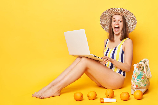 Niesamowite podekscytowana kobieta freelancer noszenie jednoczęściowy strój kąpielowy pozowanie izolowane nad żółtym tle krzyczy z podniecenia pracy na laptopie podczas odpoczynku na ośrodku. - Zdjęcie, obraz