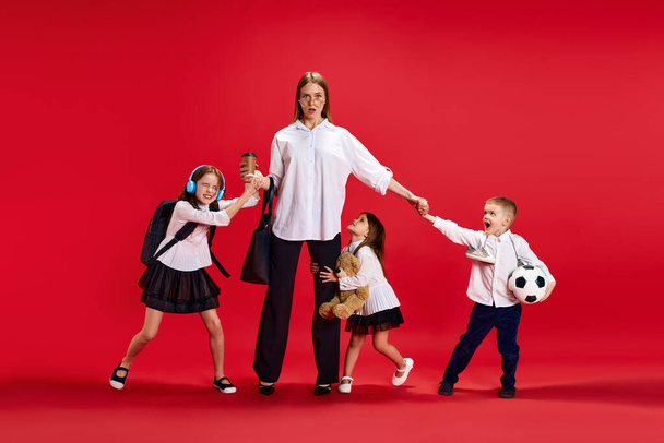 Portrait of mother and businesswoman being disturbed by children, boy and girls against red studio background. Concept of family, motherhood, childhood, fashion, lifestyle, business - Fotó, kép