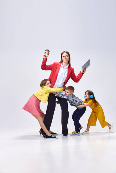 Portrait of busy woman with tablet and coffee against grey studio background. Little children hugging her, disturbing from work. Concept of family, motherhood, childhood, fashion, lifestyle - Foto, afbeelding