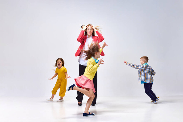 Portrait of tired young woman, mother holding head with hands around little noisy kids playing against grey studio background. Concept of family, motherhood, childhood, fashion, lifestyle - Foto, immagini