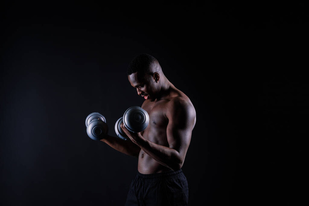 暗いスタジオの背景にダンベルを持つ孤立したアフリカの筋肉の男。強い白人黒人 - 写真・画像