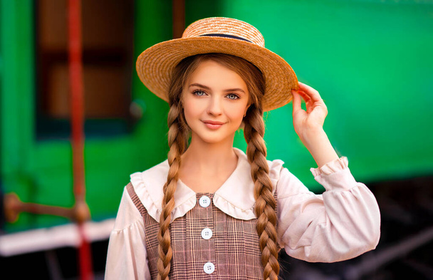 Portrait of young pretty blonde braided girl in vintage dress and straw hat standing on station platform near green train. Smiling elegance cute lady with blue eyes posing. Art work about traveller. - Foto, afbeelding