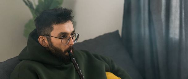 Bearded millennial or gen z man smoking hookah while relaxing on sofa at home - chill time and resting - Valokuva, kuva