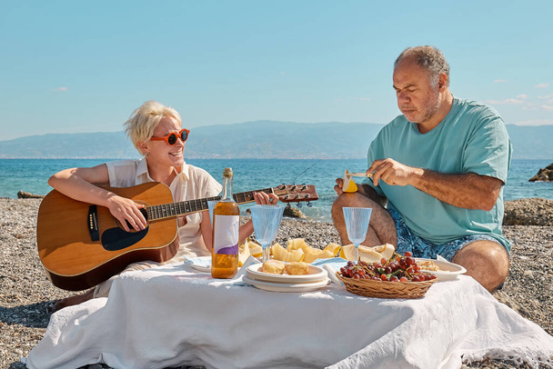 Yaz pikniği. Orta yaşlı bir çift sahilde piknik yapıyor. Sakallı adam deniz kenarında piknik yapmak için taze meyve ve aperatifler hazırlarken sarışın kadın gitar çalıyor.. - Fotoğraf, Görsel