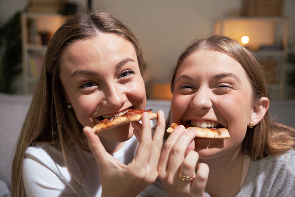 Two blonde women eating pizza and looking at camera. Friends or sisters having fun at night. - 写真・画像
