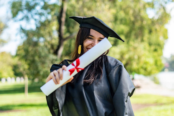Szczęśliwa biała absolwentka z długimi brązowymi włosami, pokazująca dyplom. Ma na sobie suknię kawalerską i czarną moździerz.. - Zdjęcie, obraz