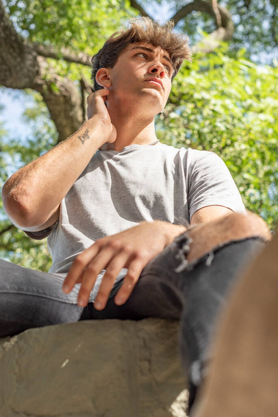 Χαμηλή οπτική γωνία ενός νεαρού άνδρα, σοβαρός, κάθεται σε ένα δέντρο, ενώ κοιτάζει μακριά - Φωτογραφία, εικόνα