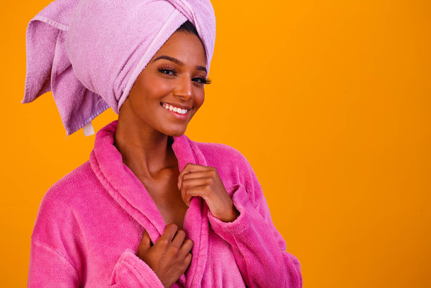 Afro woman with towel on her head and bathrobe after getting out of the bath on yellow background. Bath and care concept - Foto, Imagen