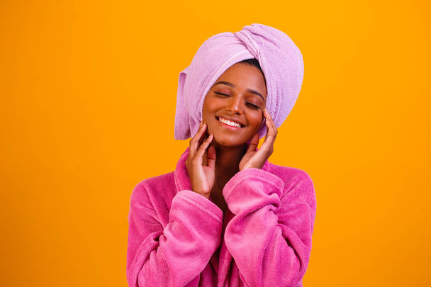 Afro woman with towel on her head and bathrobe after getting out of the bath on yellow background. Bath and care concept - Foto, afbeelding