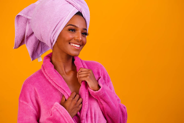 Afro woman with towel on her head and bathrobe after getting out of the bath on yellow background. Bath and care concept - Fotografie, Obrázek