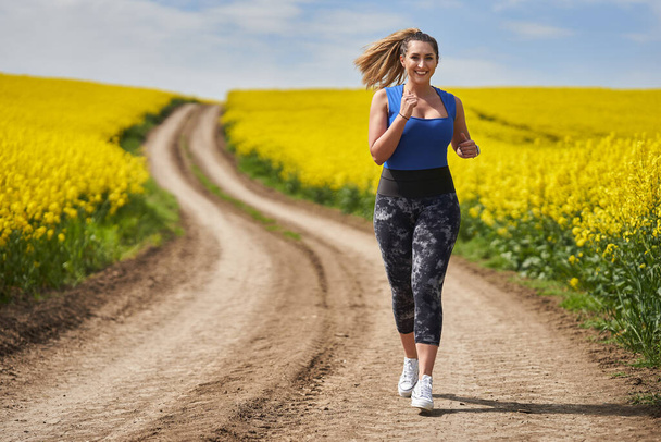 Plus size beautiful latin woman jogging on a dirt road by a canola field - Fotó, kép