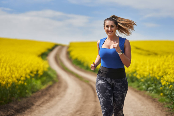 Plus size beautiful latin woman jogging on a dirt road by a canola field - Foto, Imagem