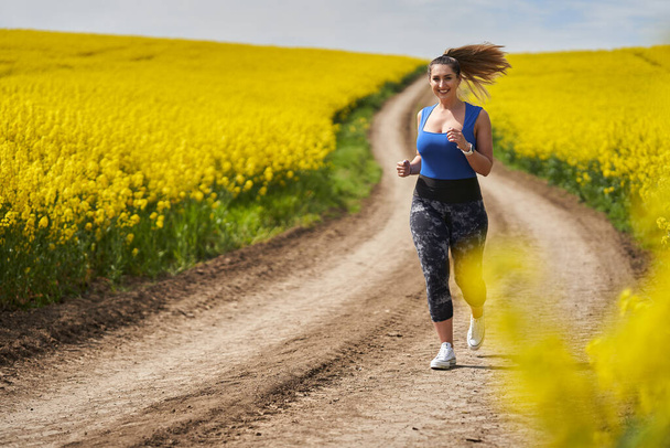 Plus size beautiful latin woman jogging on a dirt road by a canola field - Fotó, kép