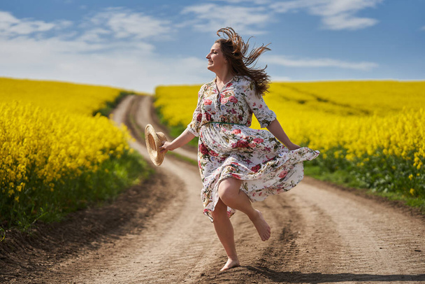 Plus grootte prachtige vrouw in een bloemenjurk, lopen en dansen blootsvoets op een onverharde weg door een bloeiende canola veld op het platteland - Foto, afbeelding