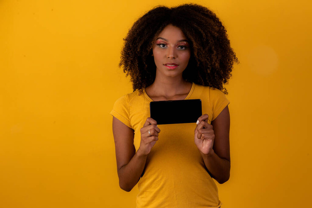 afro woman with curly hair pointing at cellphone on yellow background - Foto, immagini