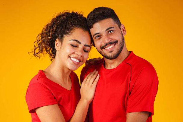 Portrait of content couple in basic clothing smiling at camera while woman putting her head on male shoulder isolated over yellow background he is surprised and happy - Valokuva, kuva