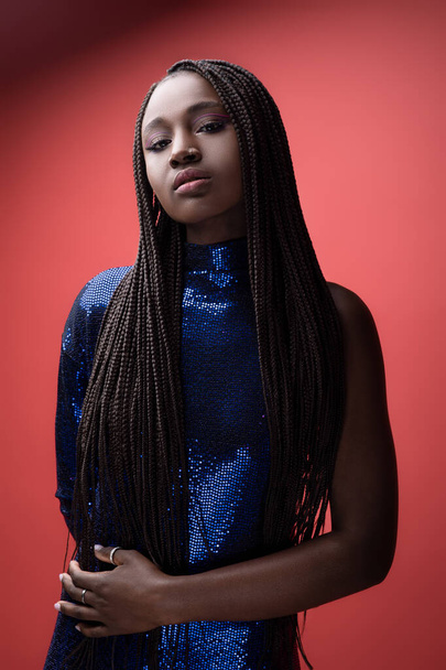 Mooie Jonge Afro-Amerikaanse vrouw dragen pailletten partij jurk poseren over Red Studio achtergrond, mode schot van zwart vrouw met artistieke make-up en gevlochten haar, stijlvolle dame op zoek naar camera - Foto, afbeelding