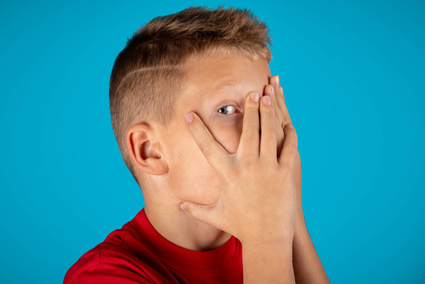 Shy preteen boy hiding face and peeking out through fingers, curious tween male child looking at camera with interest, caucasian kid posing isolated over blue studio background, copy space - Photo, Image
